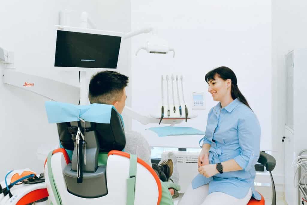 In a dental office, woman talking to a man sitting who is in the dental chair. 