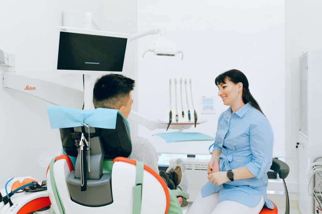 In a dental office, woman talking to a man sitting who is in the dental chair.