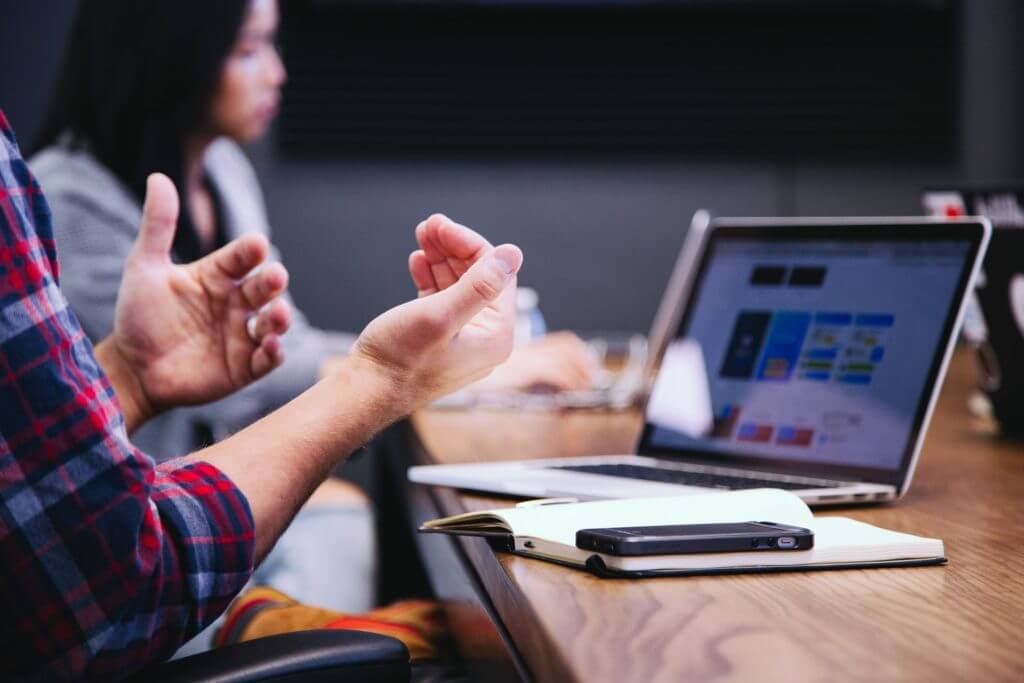 Hands gesturing next to computer screen. People gathered at a table for a meeting.