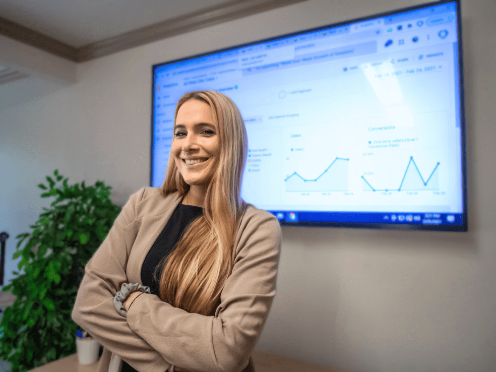 Confident blonde girl standing in front of a screen with metrics.